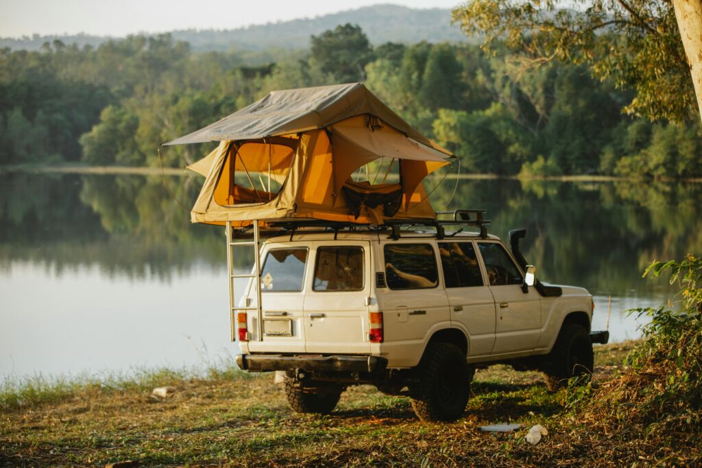 A 4x4 vehicle with a rooftop tent parked by a tranquil lake surrounded by lush forest.