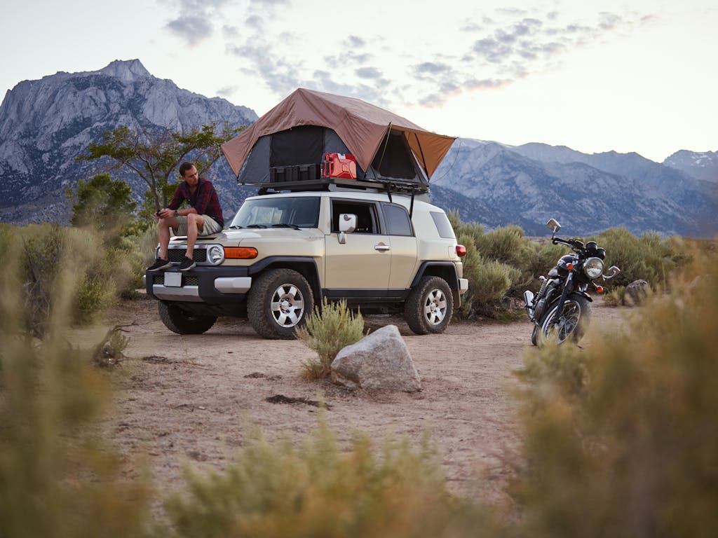 Outdoor camping scene with a man sitting on a car equipped with a rooftop tent, scenic mountain view.